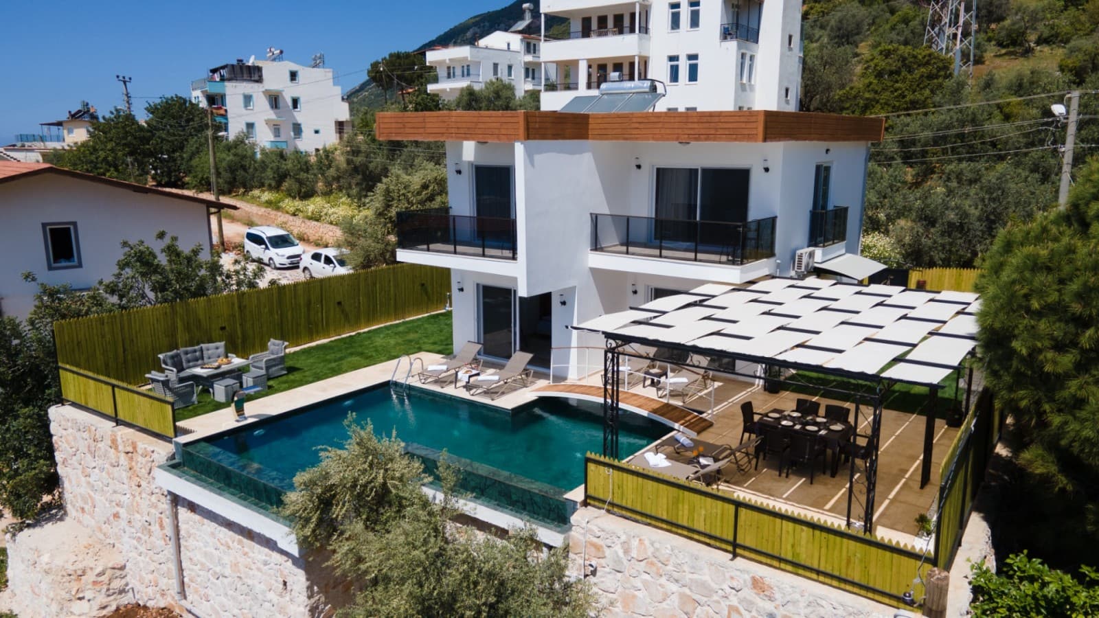 Beyaz cepheli iki katlı villanın havuzu ve pergola altındaki yemek masası.