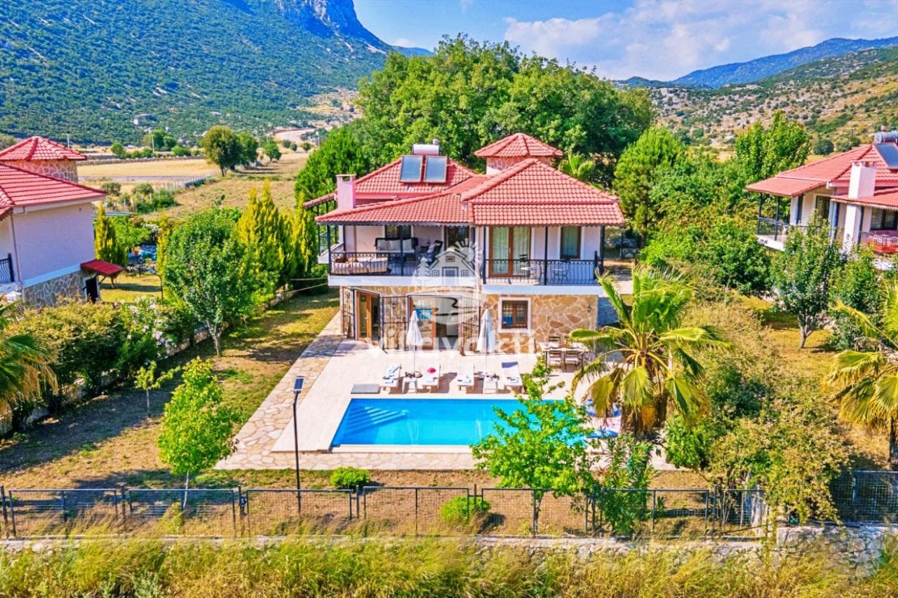 Kırmızı kiremit çatılı taş villanın önünde havuz ve bahçe manzarası görülüyor.