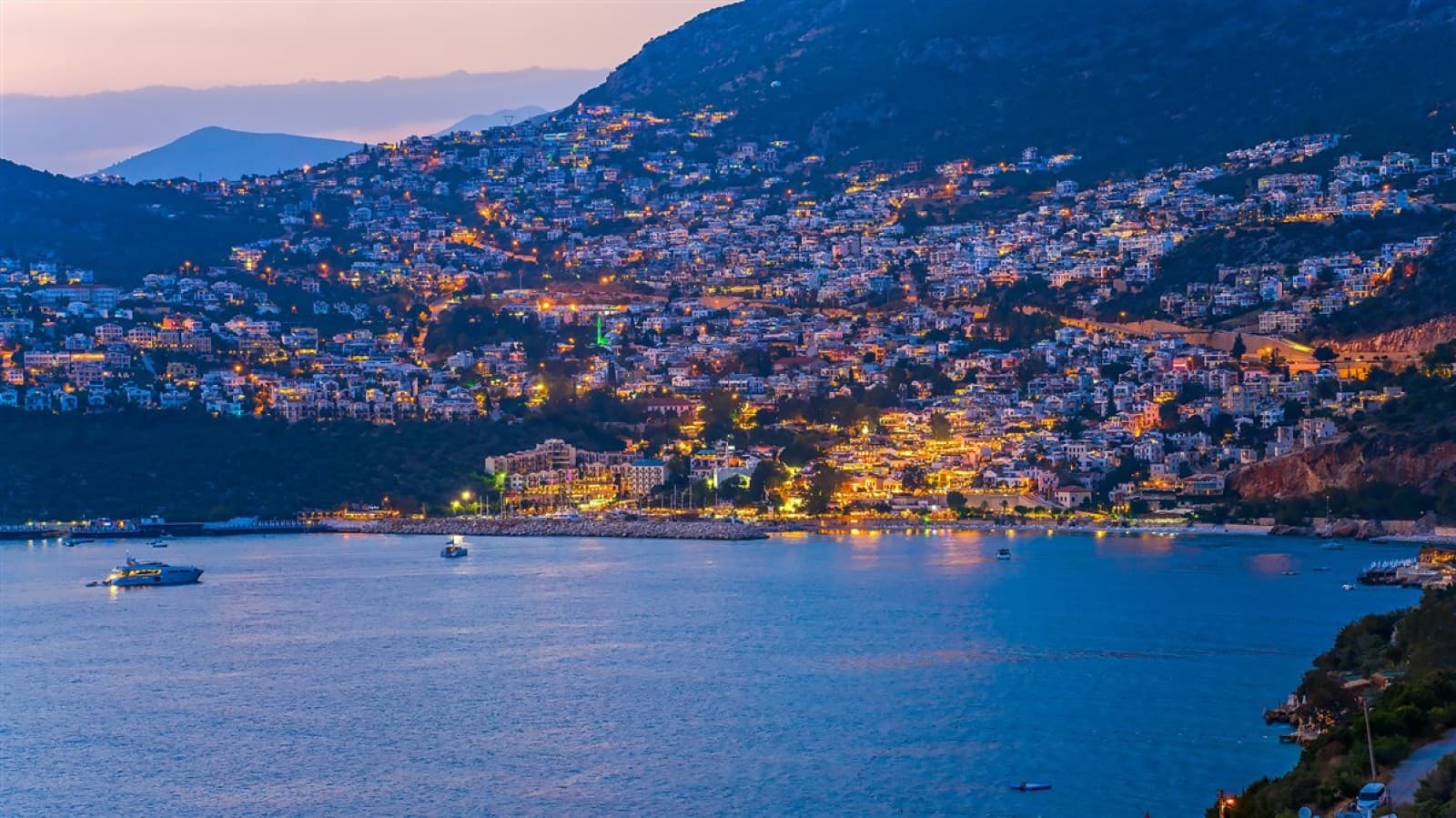Akşam saatlerinde yamaçta ışıklandırılmış sahil kasabası ve deniz manzarası.