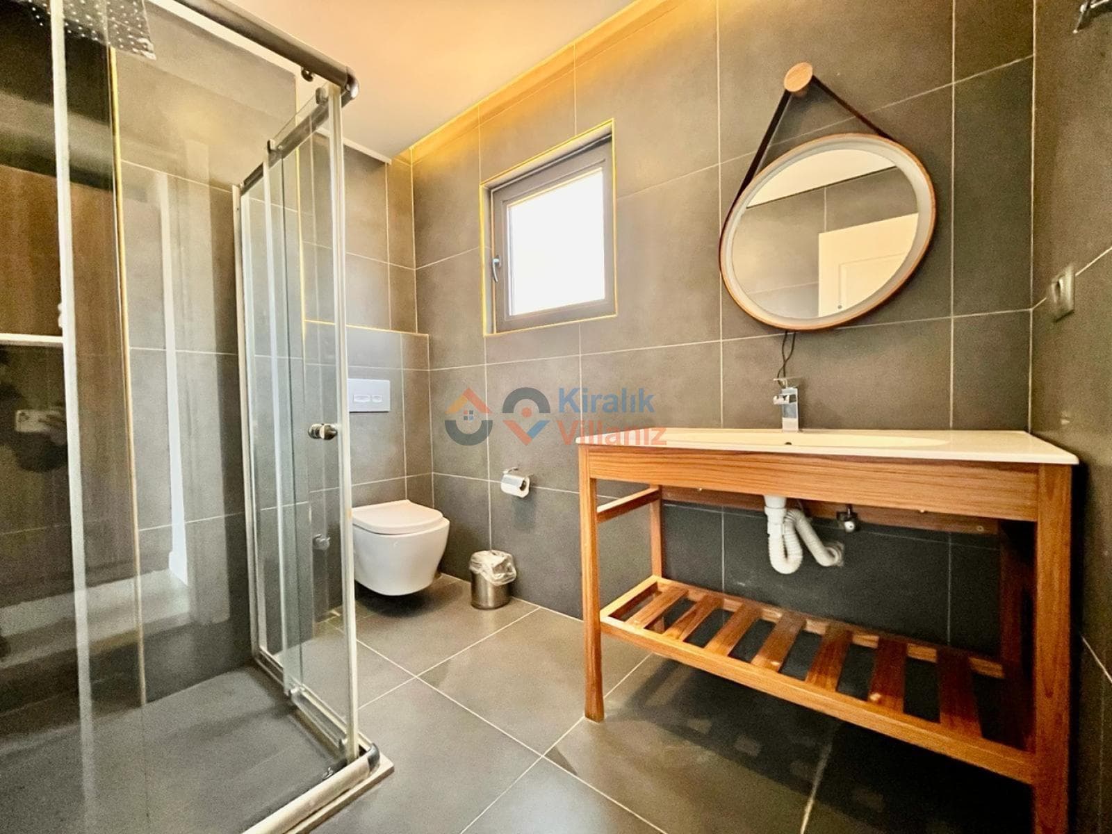 Gri fayanslı modern banyoda cam duşakabin, ahşap lavabo dolabı ve yuvarlak ayna bulunuyor.