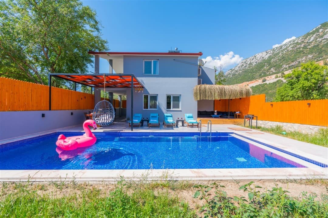 Dağ manzaralı havuzlu villanın bahçesinde şezlonglar ve pembe flamingo deniz yatağı var.