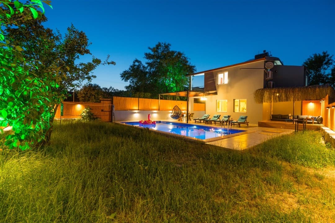 Çimenlik bahçede aydınlatılmış havuz ve iki katlı villa.