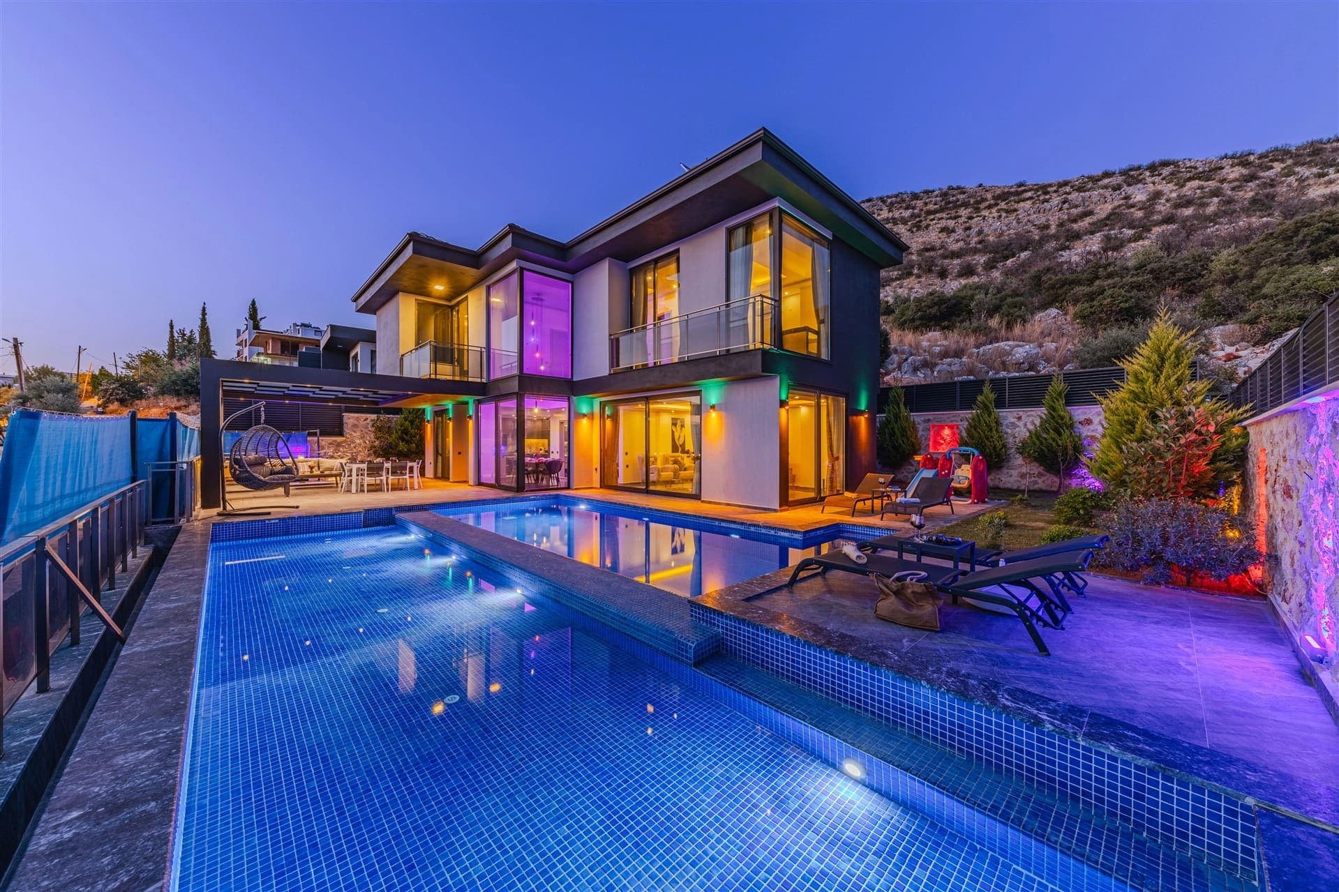 Akşam aydınlatmalı iki katlı modern villanın önünde havuz ve şezlonglar bulunuyor.