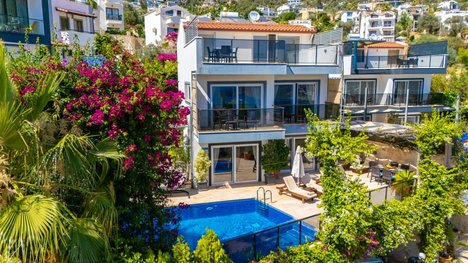 Önünde havuz bulunan, pembe sarmaşıklarla çevrili çok katlı beyaz villa dış cephesi.