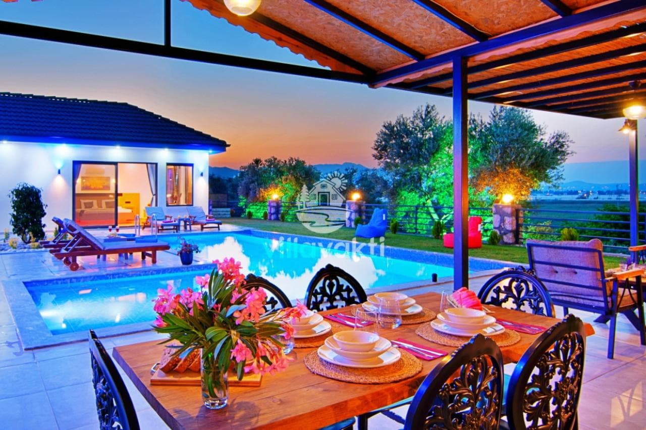 Ön planda akşam yemeği masası ve arkada havuzlu villanın akşam manzarası.