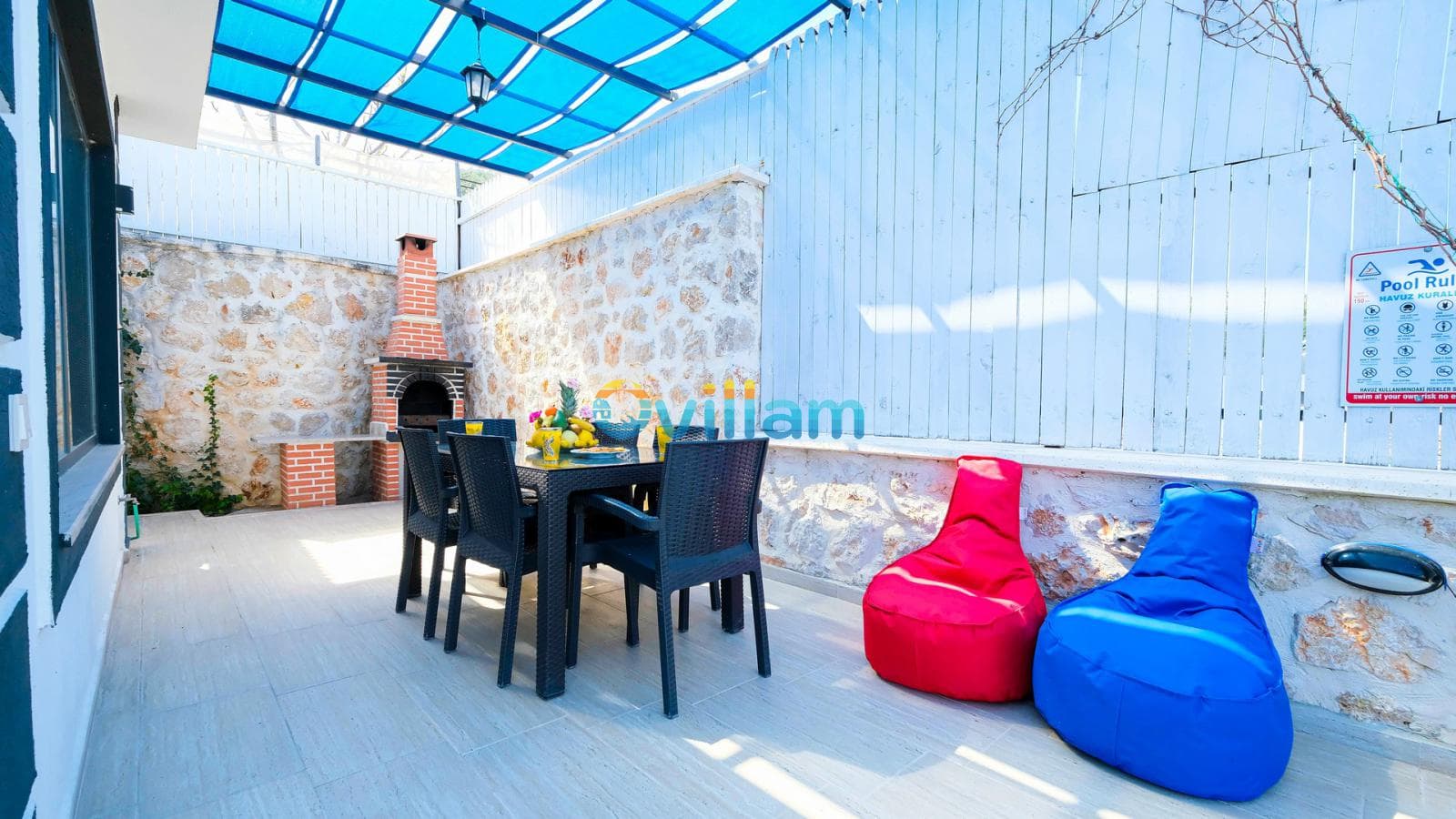 Taş duvarlı villanın terasında yemek masası, barbekü, kırmızı ve mavi minderler bulunur.