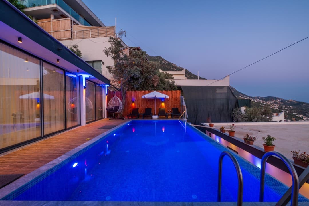 Mavi aydınlatmalı havuz kenarında şezlonglar ve cam cepheli villa yer alıyor.