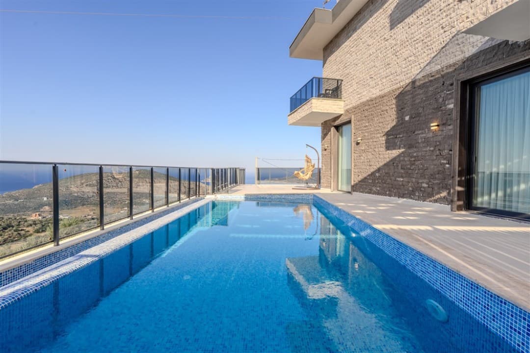 Cam korkuluklu havuz kenarında taş duvarlı villa ve dağ manzarası.