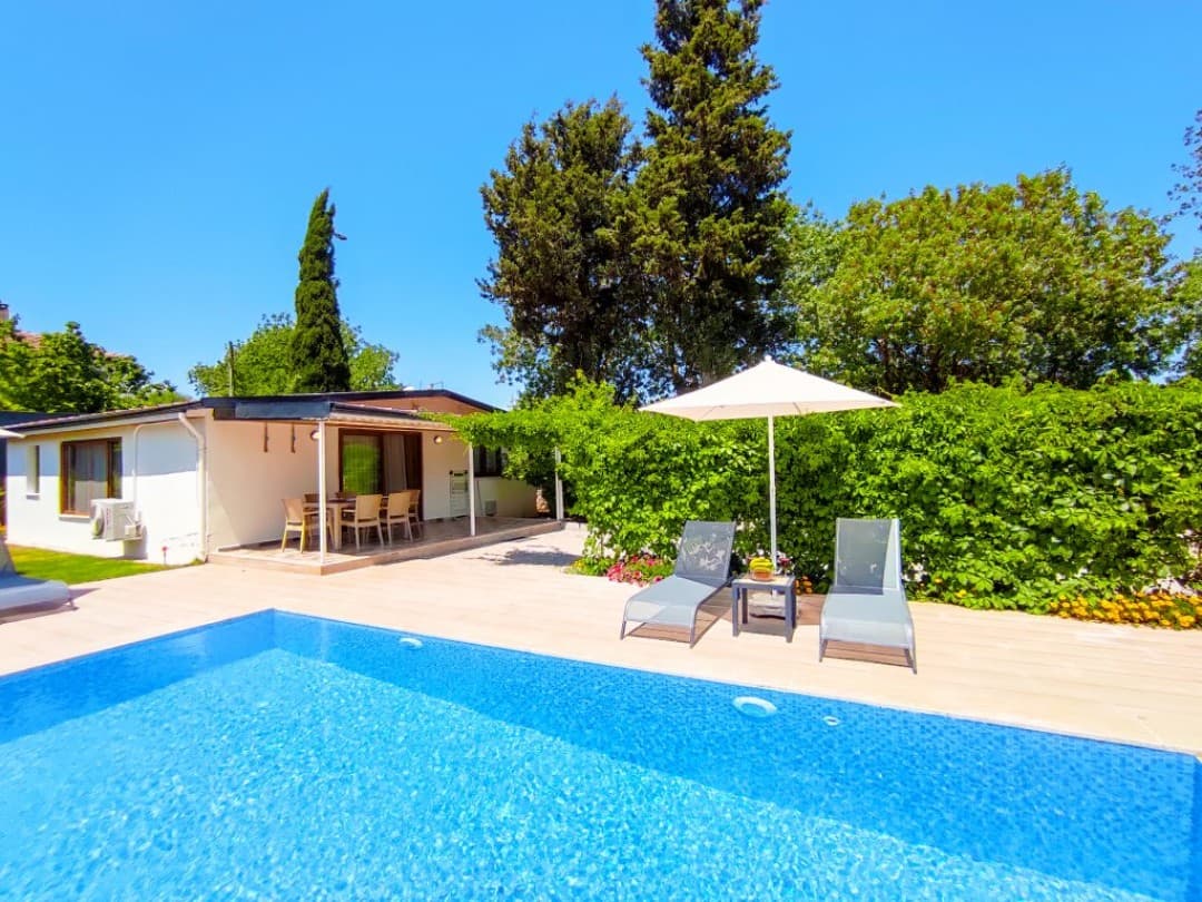Mavi yüzme havuzu, yanında şezlonglar, güneş şemsiyesi ve veranda kısmında yemek masası olan tek katlı villa.