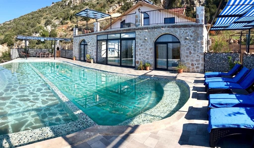 Taş kaplamalı iki katlı villanın önünde şezlonglu büyük havuz ve dağ manzarası.