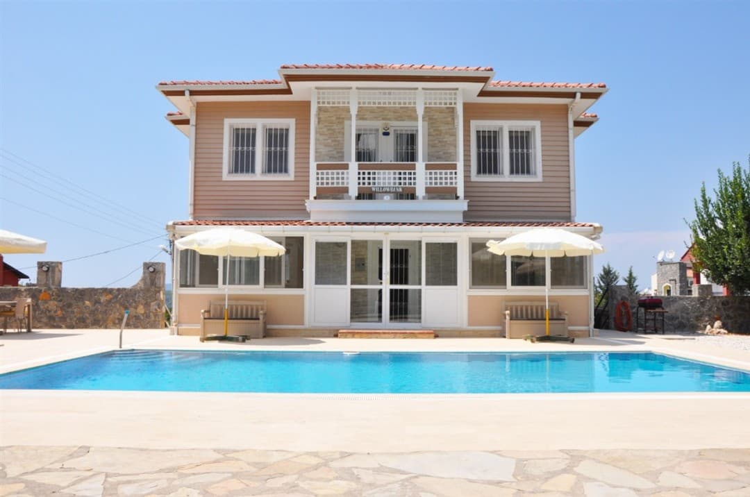 Önünde yüzme havuzu, şemsiyeler ve banklar bulunan iki katlı, ahşap görünümlü müstakil villa.