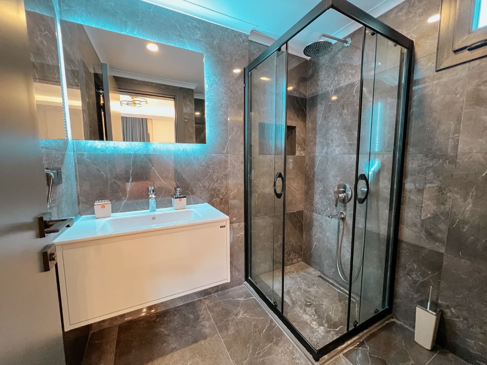 Mermer detaylı, siyah duş kabinli ve LED aydınlatmalı aynalı banyo.