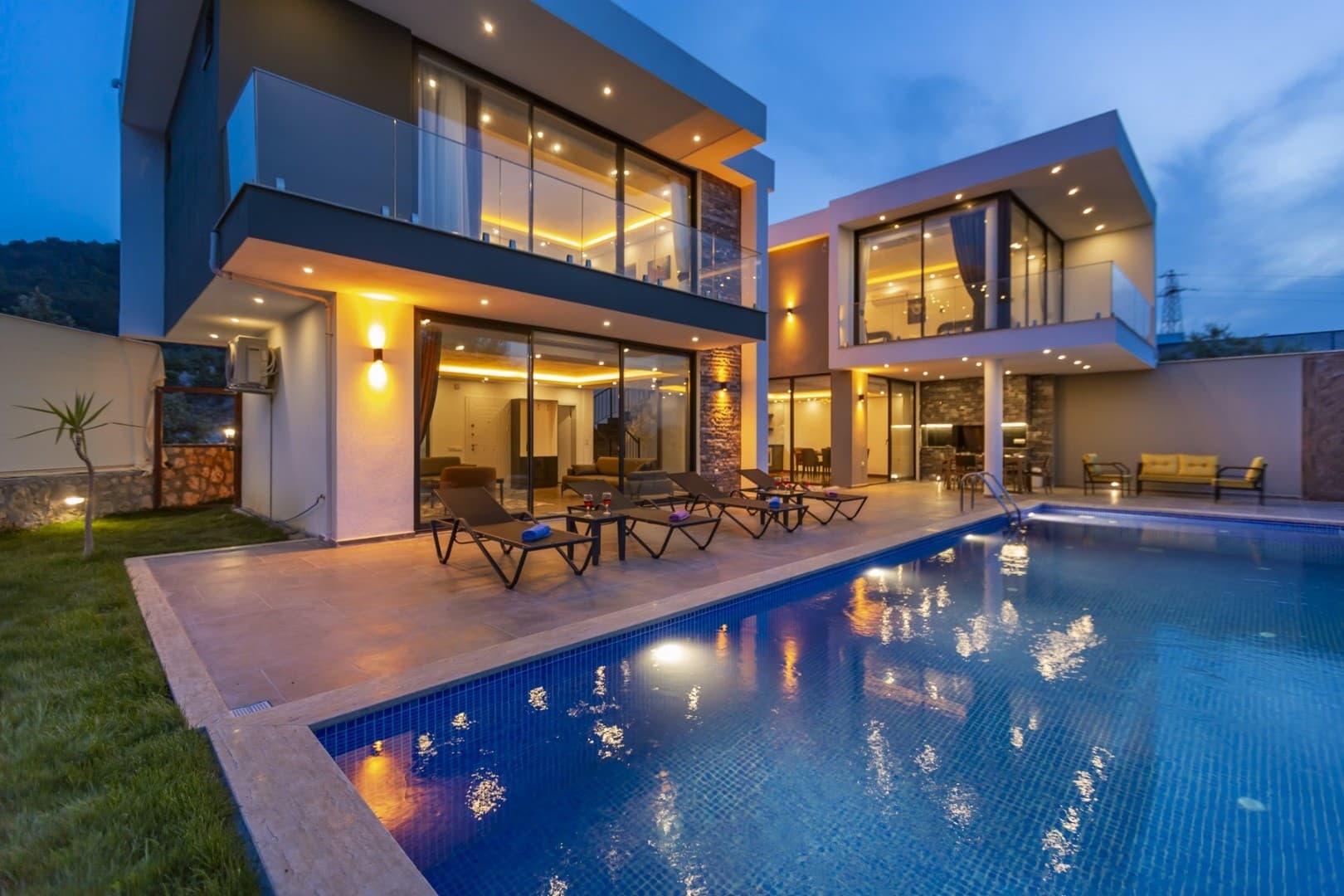 Akşam vakti aydınlatılmış, havuz başında şezlongları bulunan modern mimarili iki katlı villa.