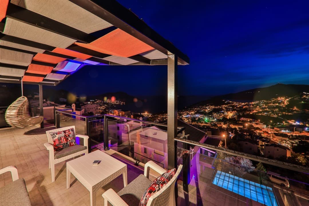 Gece vakti şehir ve deniz manzaralı balkonda beyaz masa, sandalyeler ve salıncak var.