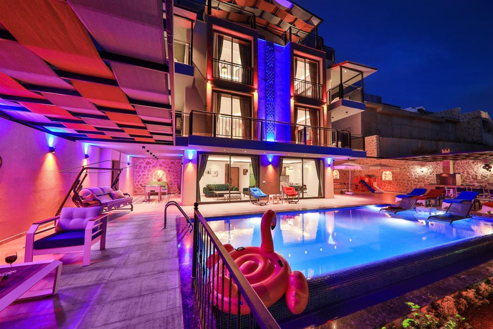 Gece aydınlatmalı havuz başı, şezlonglar ve pembe flamingo simidi olan çok katlı villa dış cephesi.