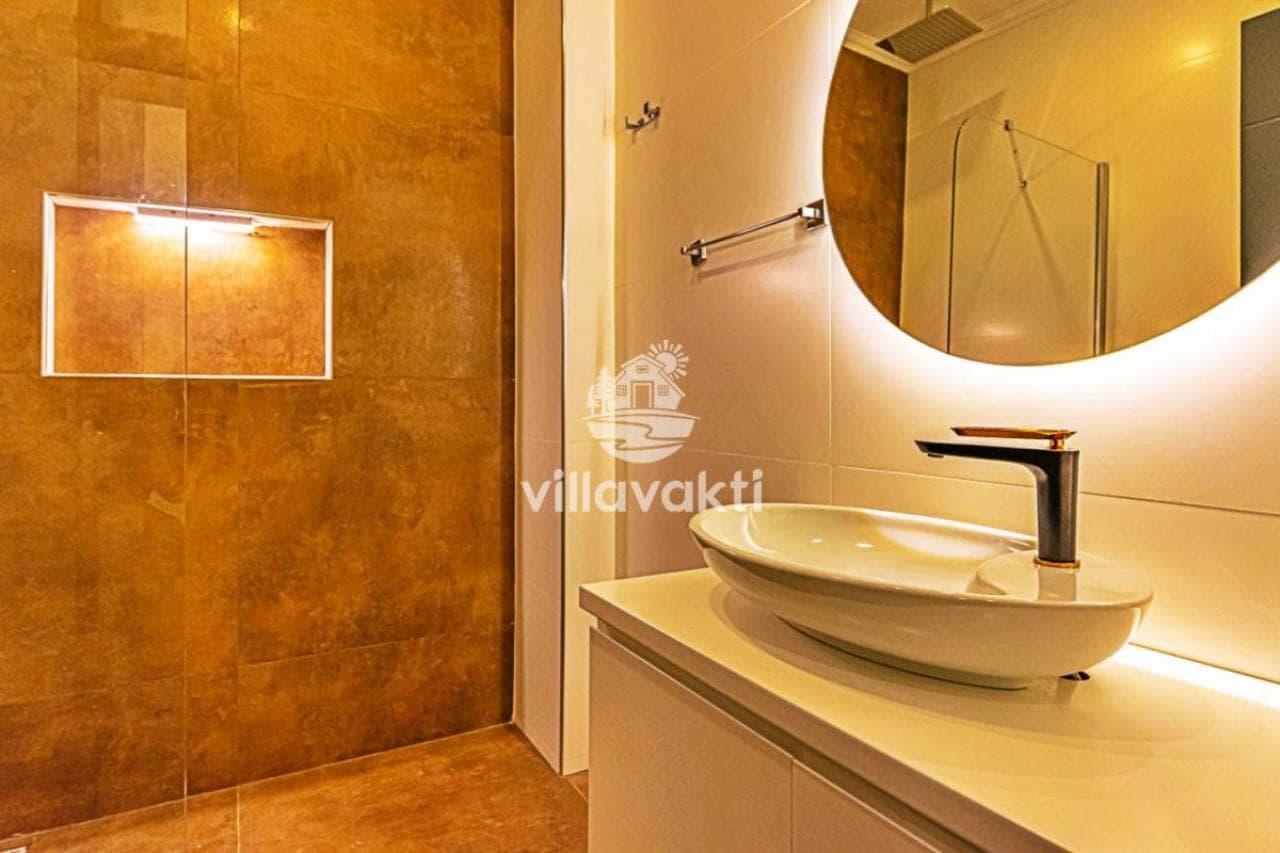 Banyoda modern lavabo, yuvarlak ayna ve duş alanı.