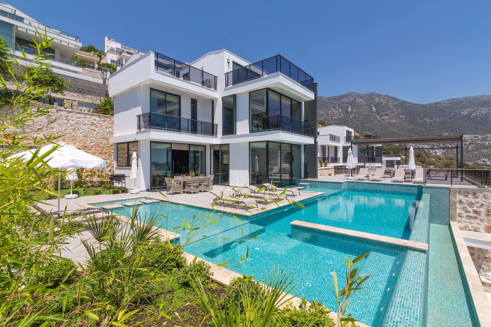 Dağlar önünde konumlanan, şezlonglu ve ferah teraslı beyaz dış cepheli villa.