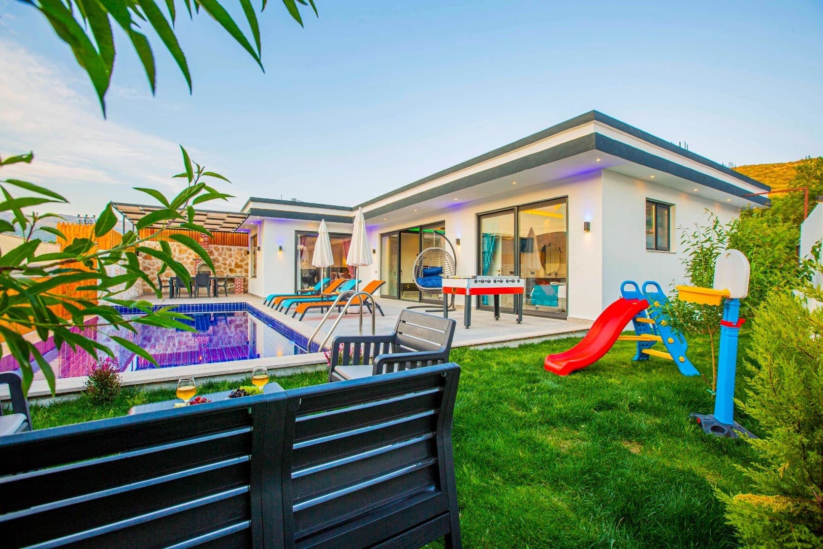 Bahçesinde havuz, çocuk oyun alanı ve oturma grubu bulunan modern villa dış cephesi.
