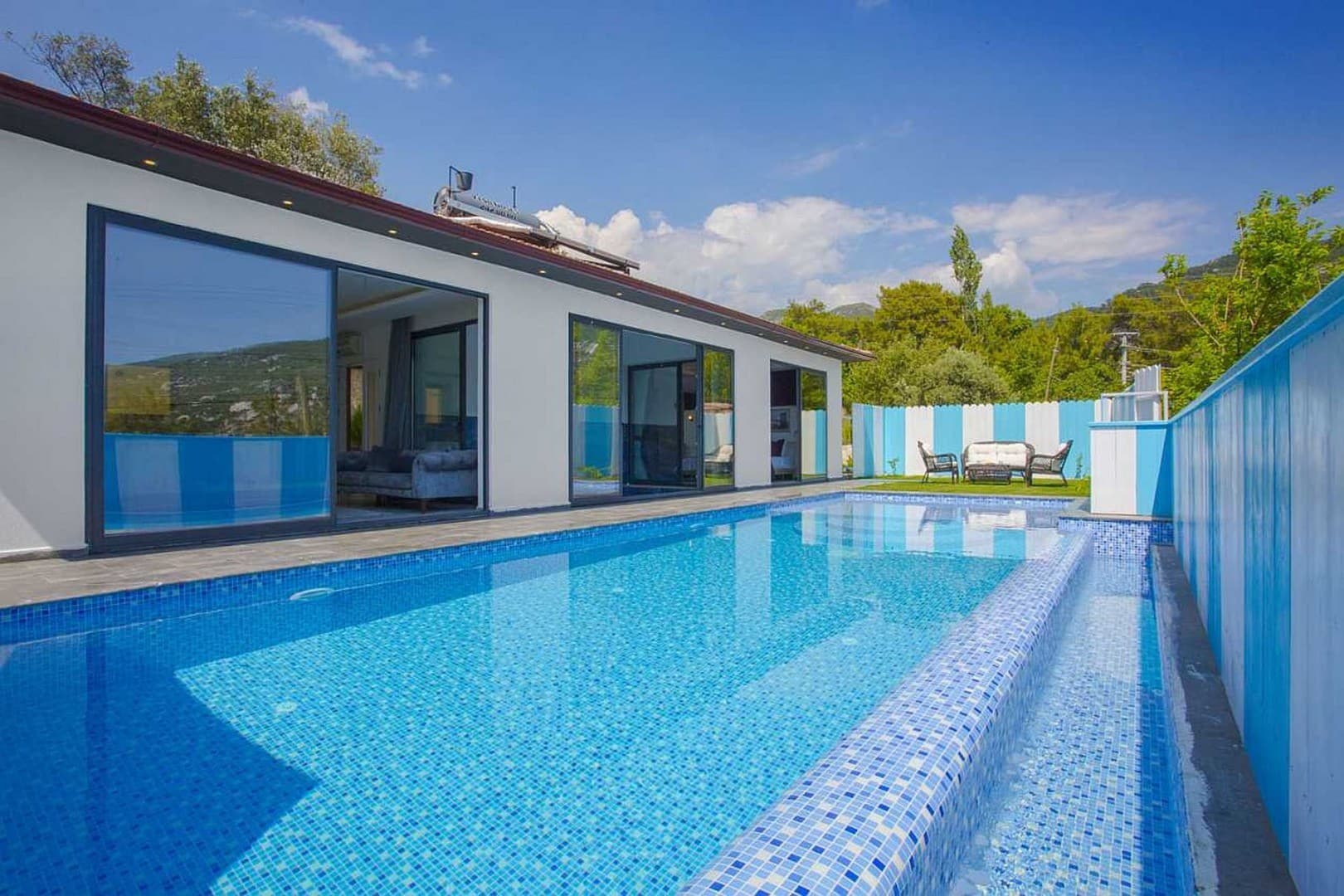 Mavi beyaz çizgili duvarlarla çevrili bahçesinde özel yüzme havuzu bulunan tek katlı modern villa.