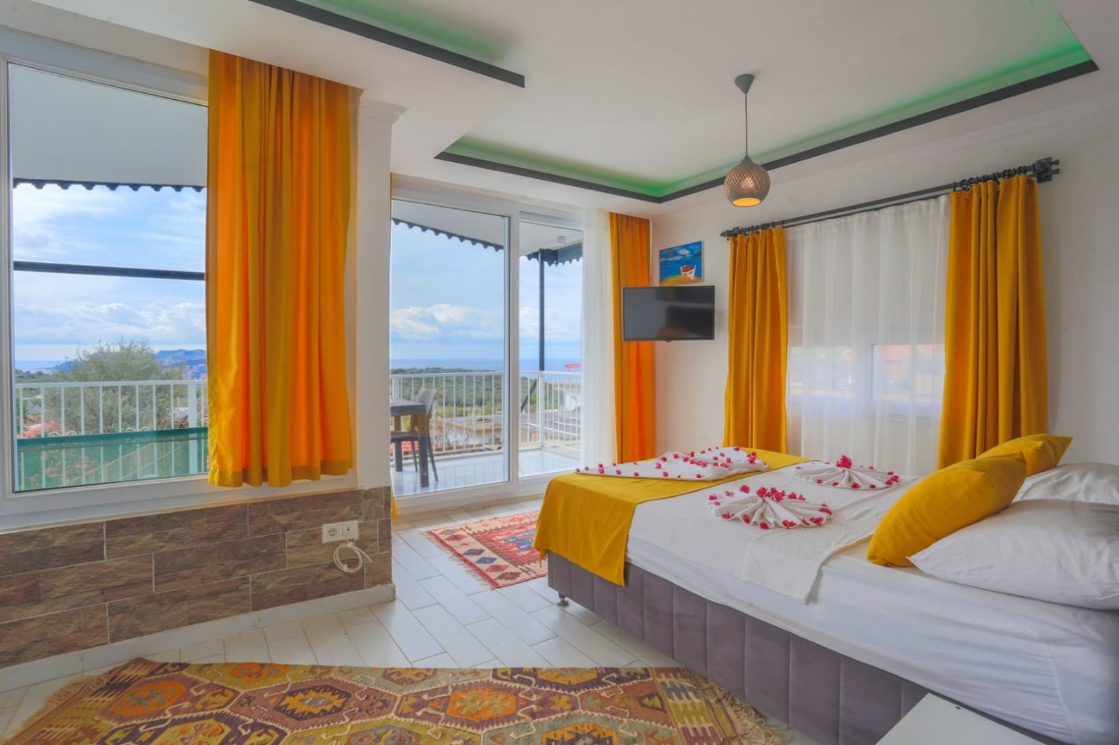 Sarı perdeli ve deniz manzaralı balkona açılan pencereli yatak odası.