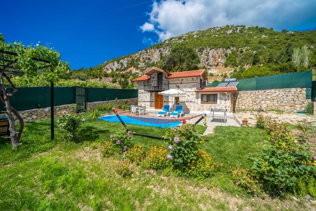 Dağ eteğinde, önünde havuz, şezlonglar ve hamak bulunan taş duvarlı müstakil ev.