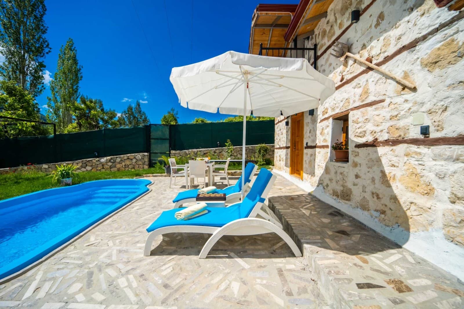 Taş duvarlı villa önünde havuz, şezlonglar ve beyaz şemsiye.