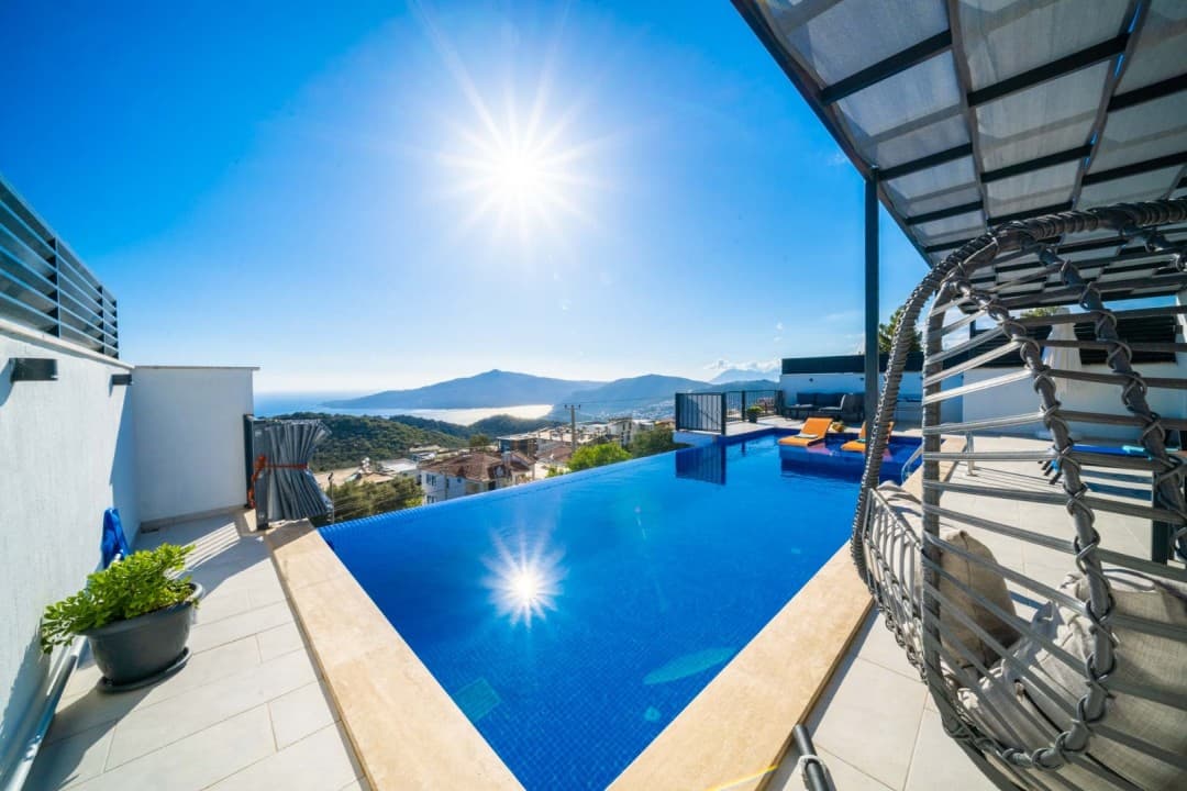 Güneşli bir günde deniz ve dağ manzaralı sonsuzluk havuzu, şezlonglar ve asma salıncak bulunan villa terası.