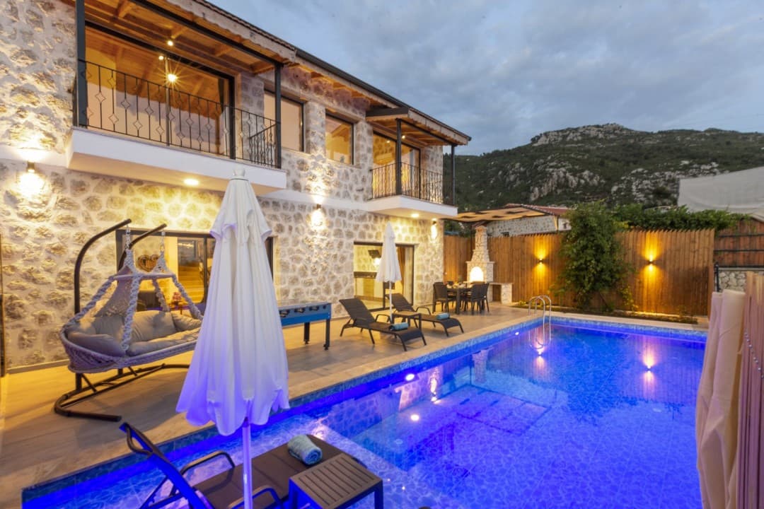 Akşam ışıklarıyla aydınlatılmış taş villa, özel havuz, şezlonglar, salıncak ve bahçe mobilyaları.