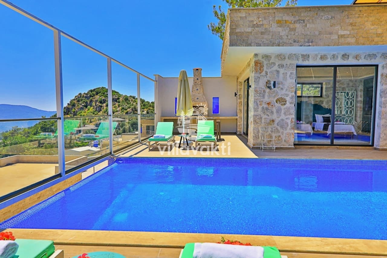 Mavi yüzme havuzu ve şezlongların bulunduğu taş villa terası ve dağ manzarası.