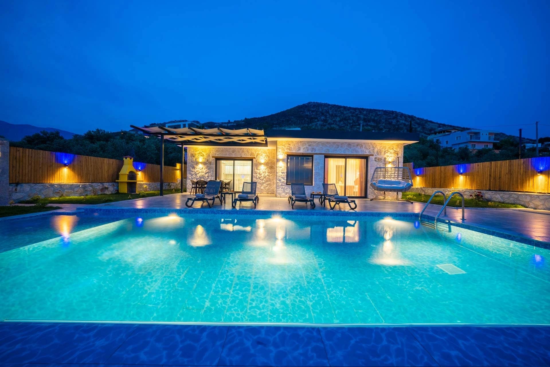 Akşam ışıklandırmalı havuz ve arkasında dağ manzaralı villa dış cephesi.