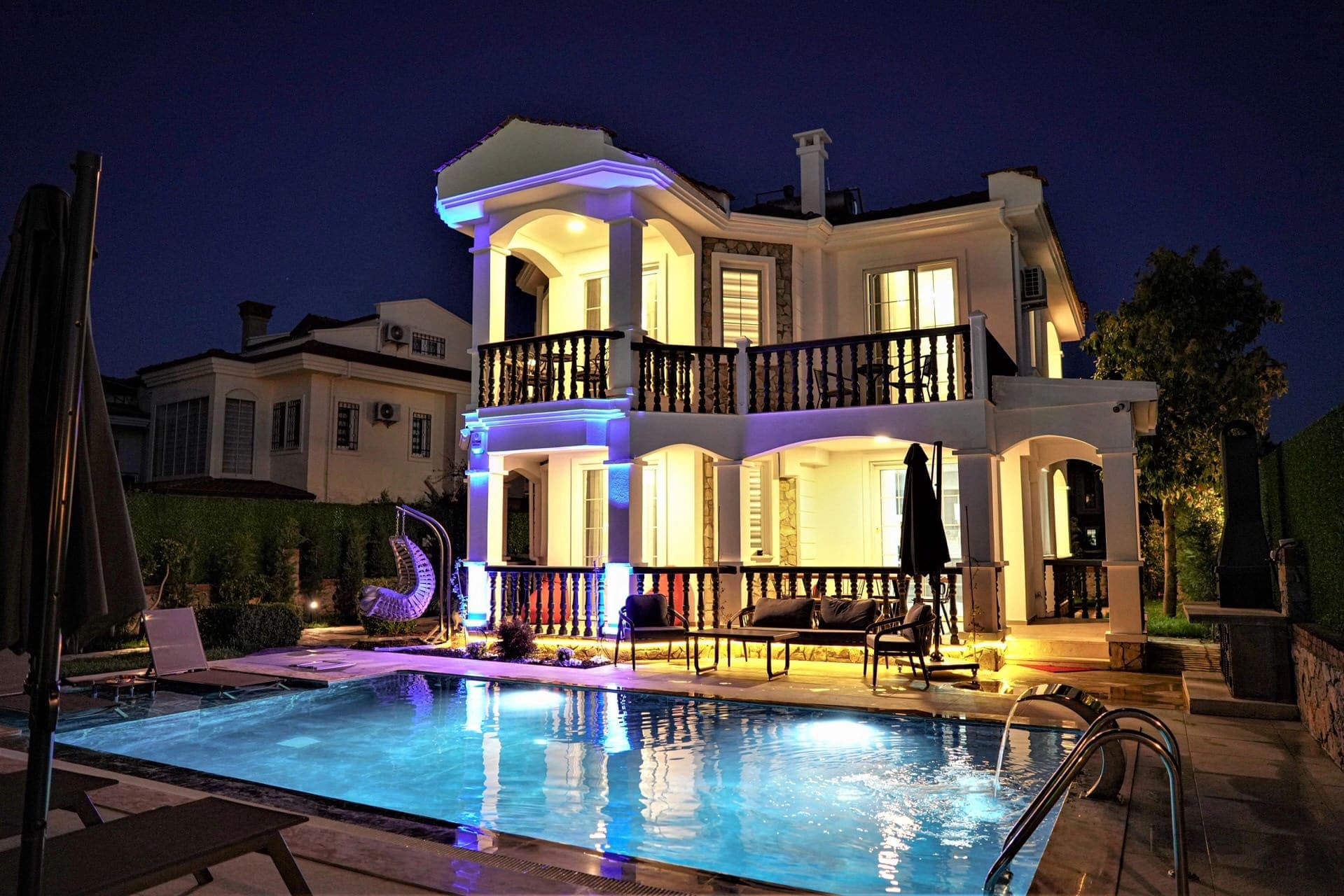 Gece aydınlatmalı havuz kenarında iki katlı beyaz villa.