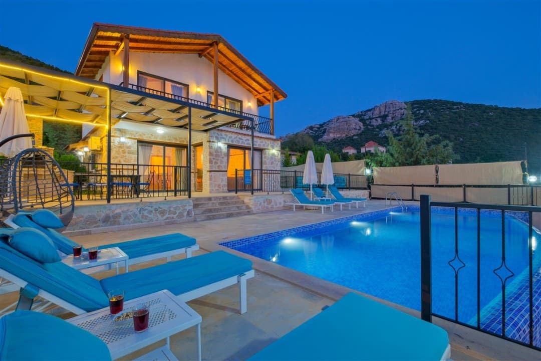 Akşam vakti aydınlatılmış taş villa, özel havuz, şezlonglar ve arka planda dağ manzarası.