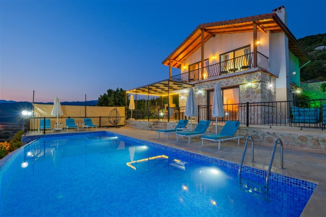 Akşam ışıklandırılmış iki katlı taş villa ve önünde aydınlatmalı havuz.