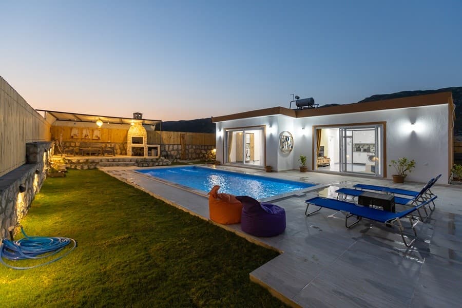 Akşam vakti havuzlu, bahçesinde şezlonglar, armut koltuklar ve taş barbekü bulunan tek katlı villa.