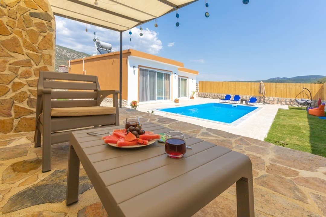 Ön planda sehpa üzerinde karpuz dilimleri ve içecekler, arka planda havuz ve villa dış cephesi.