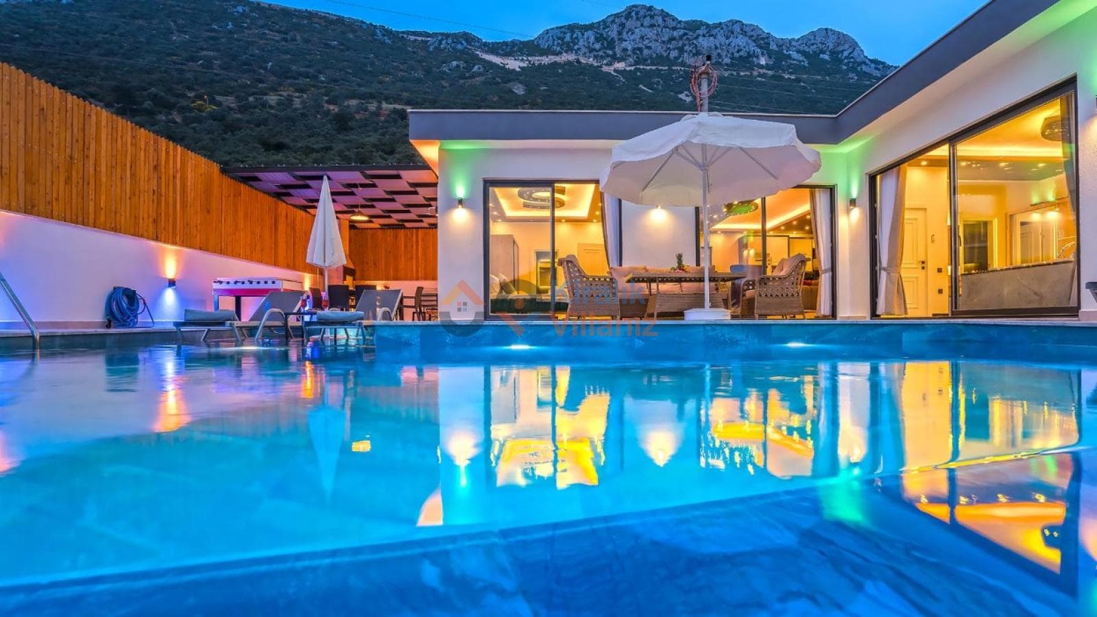 Akşamüstü aydınlatmalı havuz ve arka planda dağ manzaralı villa dış cephesi.