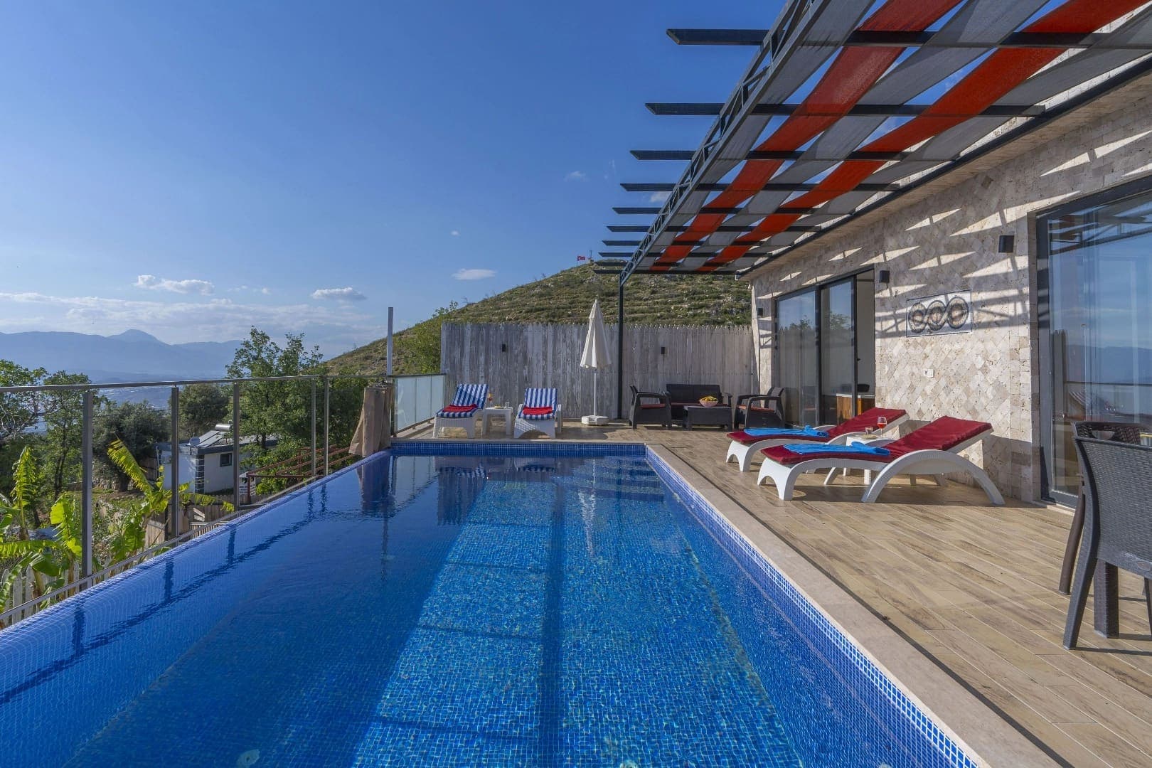 Ahşap teraslı, şezlonglu havuz başı ve uzakta dağ manzarası