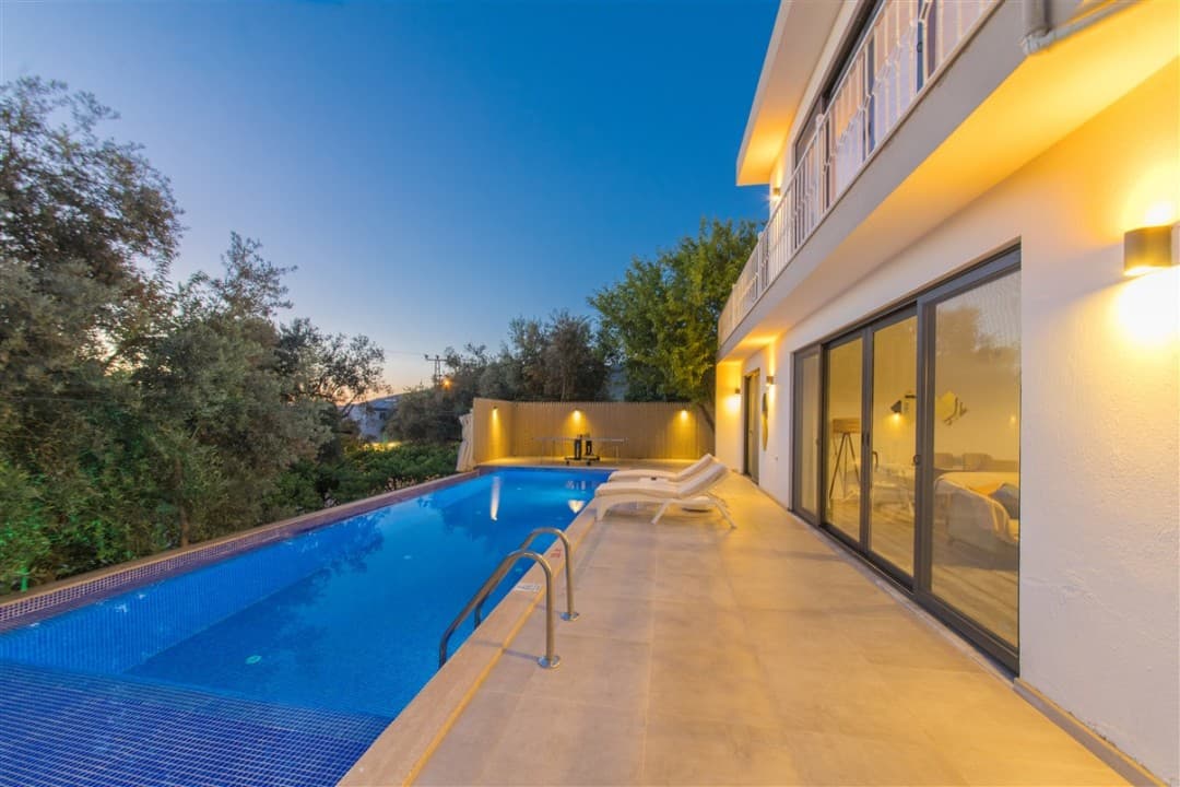 Akşam vakti aydınlatmalı havuz kenarında şezlonglar bulunan iki katlı villa.
