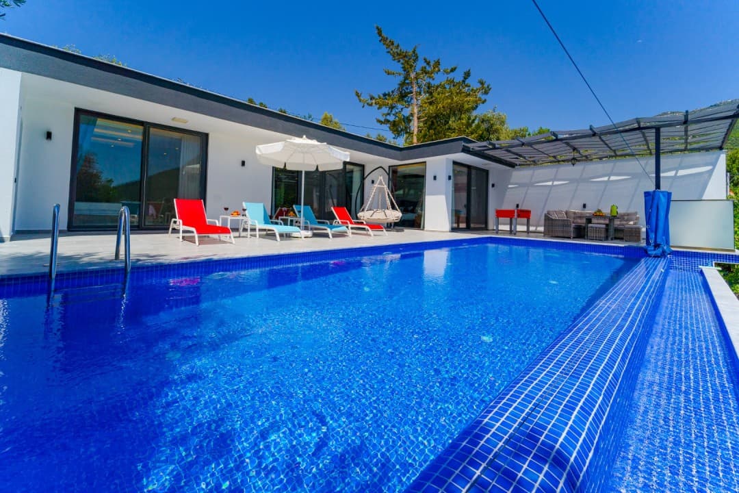 Ön planda mavi havuz, havuz kenarında renkli şezlonglar ve şemsiye bulunan tek katlı modern villa.