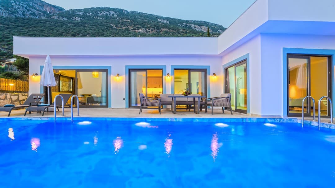 Aydınlatmalı havuz kenarında masa takımı bulunan beyaz villa dış cephesi.