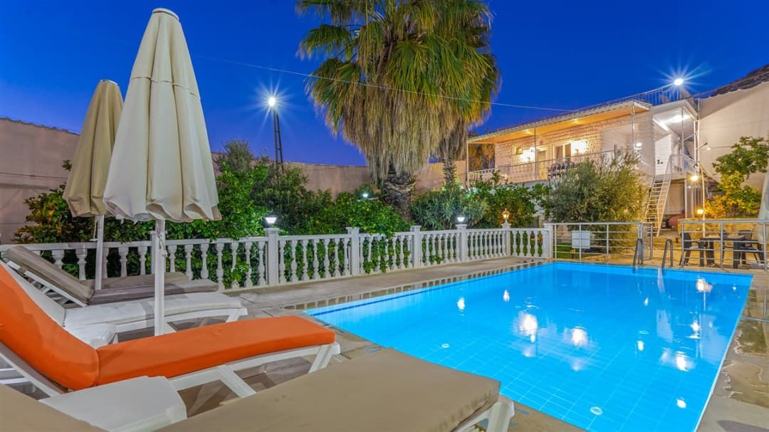 Akşam karanlığında aydınlatılmış havuz başında şezlonglar ve arka planda iki katlı villa.