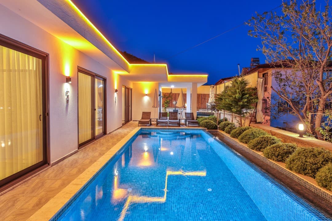 Gece çekiminde sarı ışıklandırmalı uzun havuzu ve şezlongları olan modern villa cephesi.