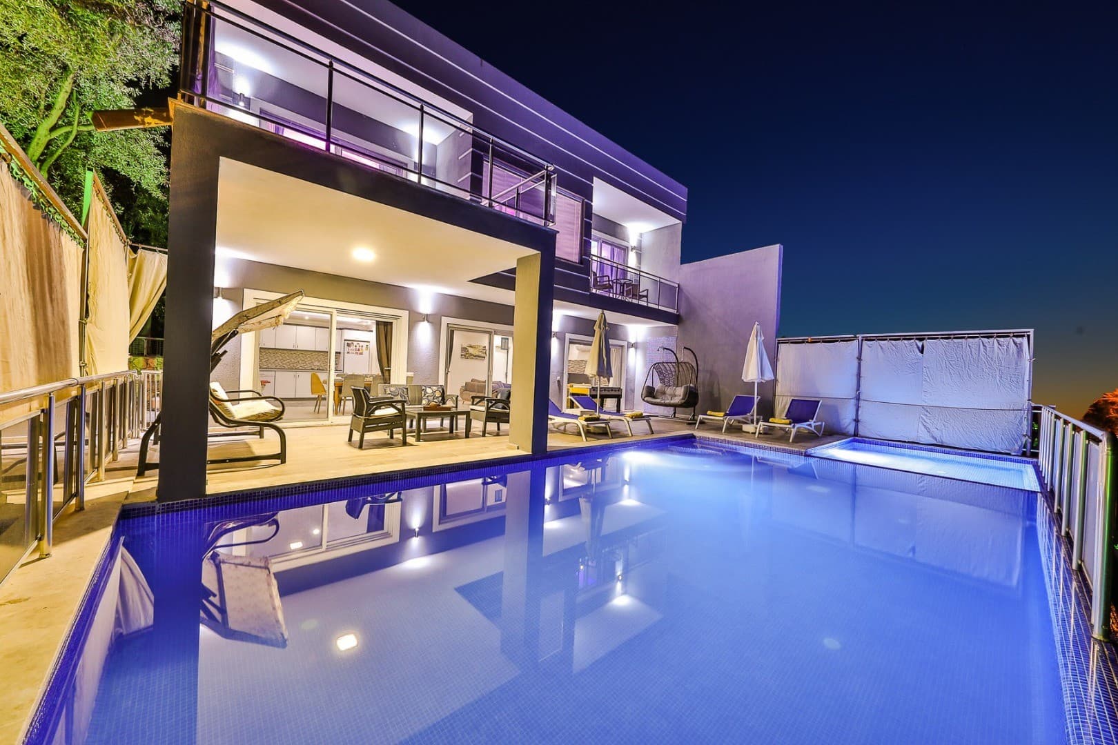 Gece aydınlatmalı havuzu, şezlongları ve oturma grubu bulunan modern iki katlı villa.