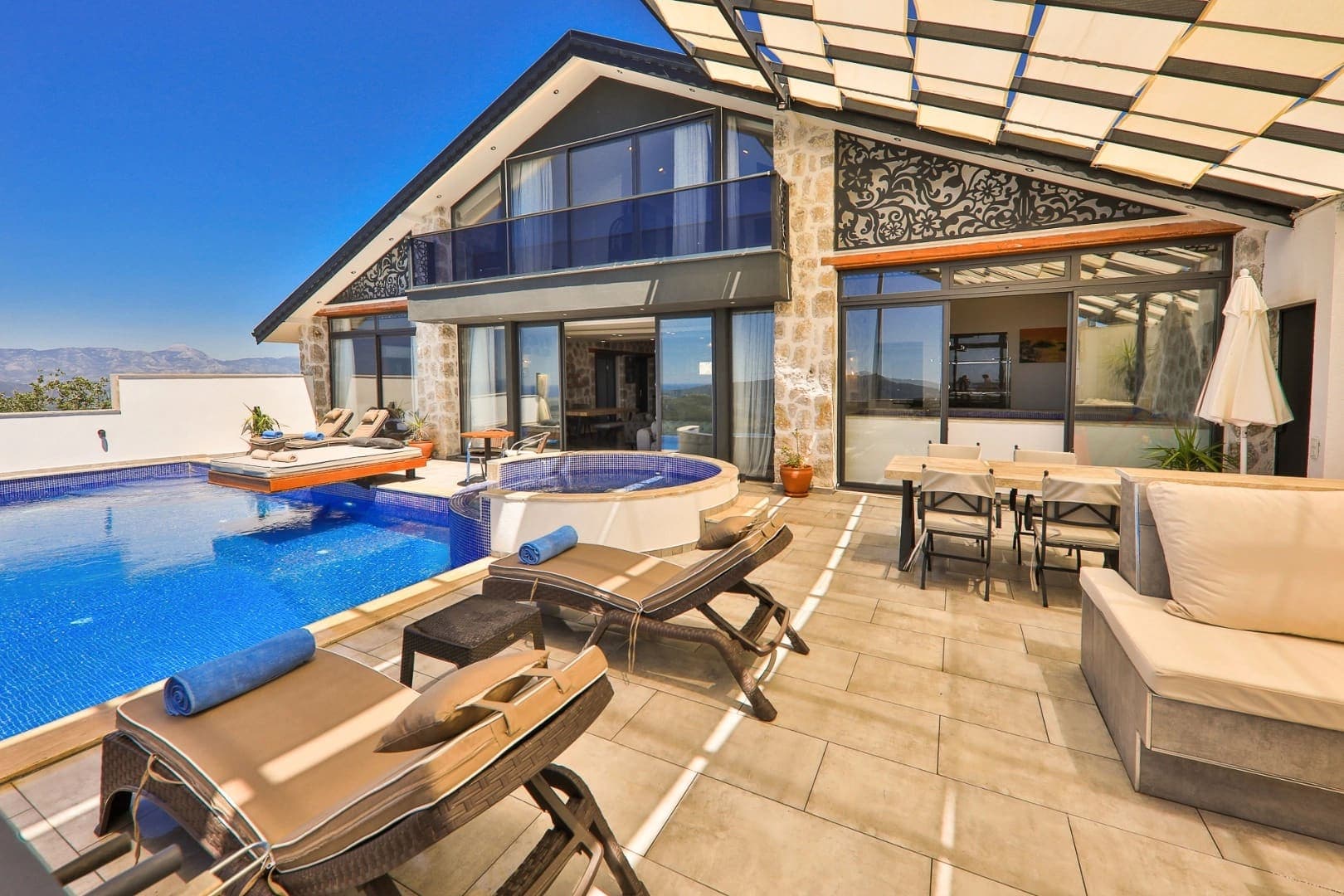 Güneşli günde villa havuzu, şezlonglar, jakuzi ve yemek masası.