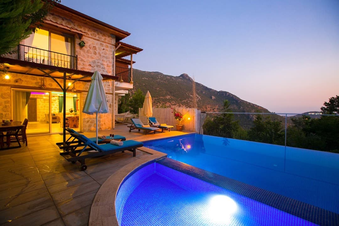 Akşam vakti havuz başı şezlongları, taş duvarlı iki katlı villa ve dağ manzarası.