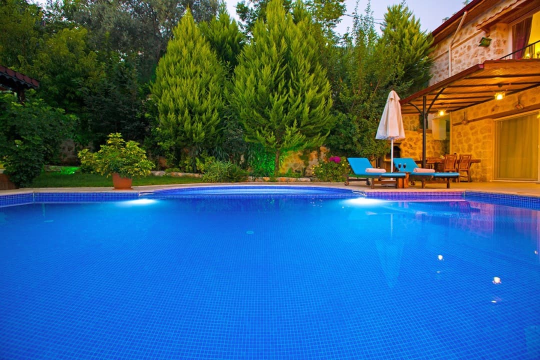 Yemyeşil ağaçlar arasında yer alan açık havuz ve taş duvarlı villa cephesi.