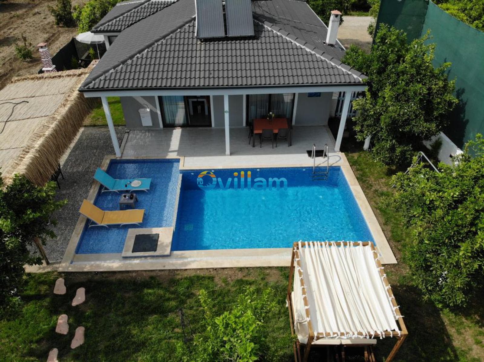 Havuz kenarında şezlonglar, pergola ve yeşillikler bulunan villanın havadan görünümü.