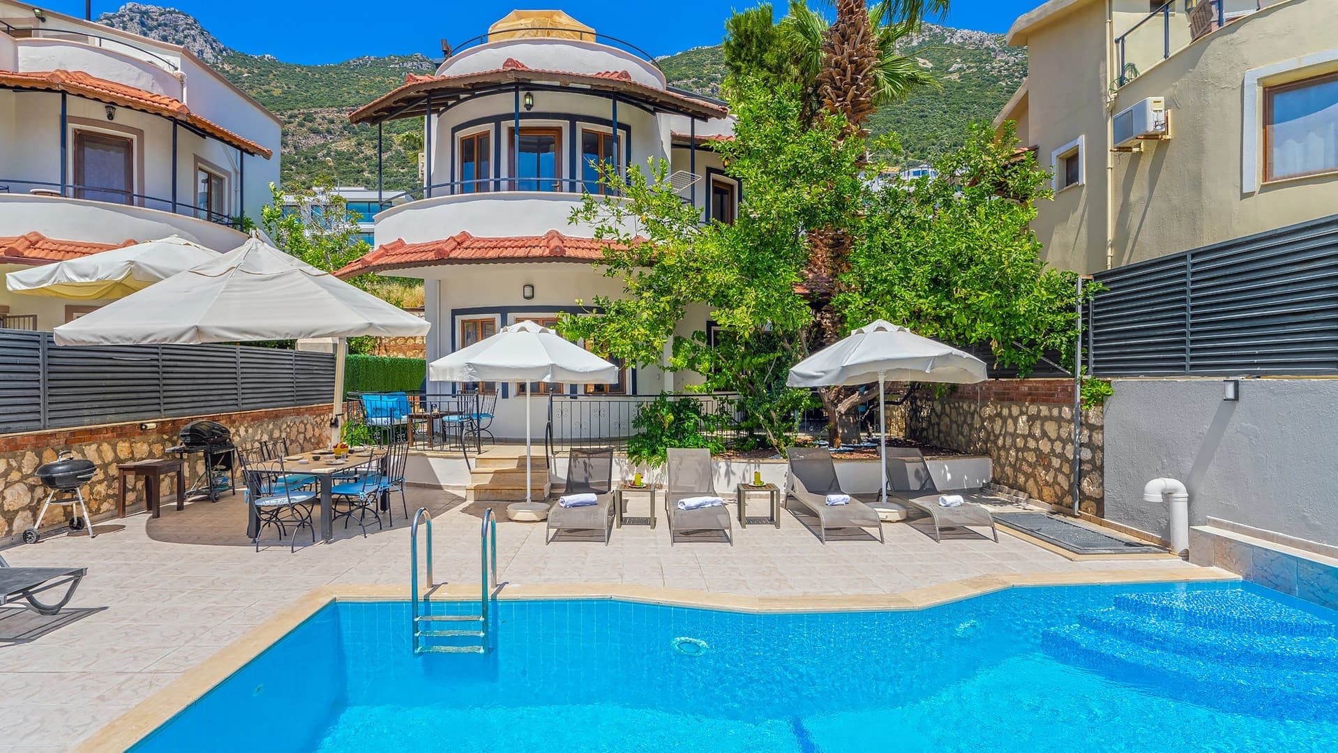 Mavi yüzme havuzu, şezlonglar, güneş şemsiyeleri ve iki katlı villa.