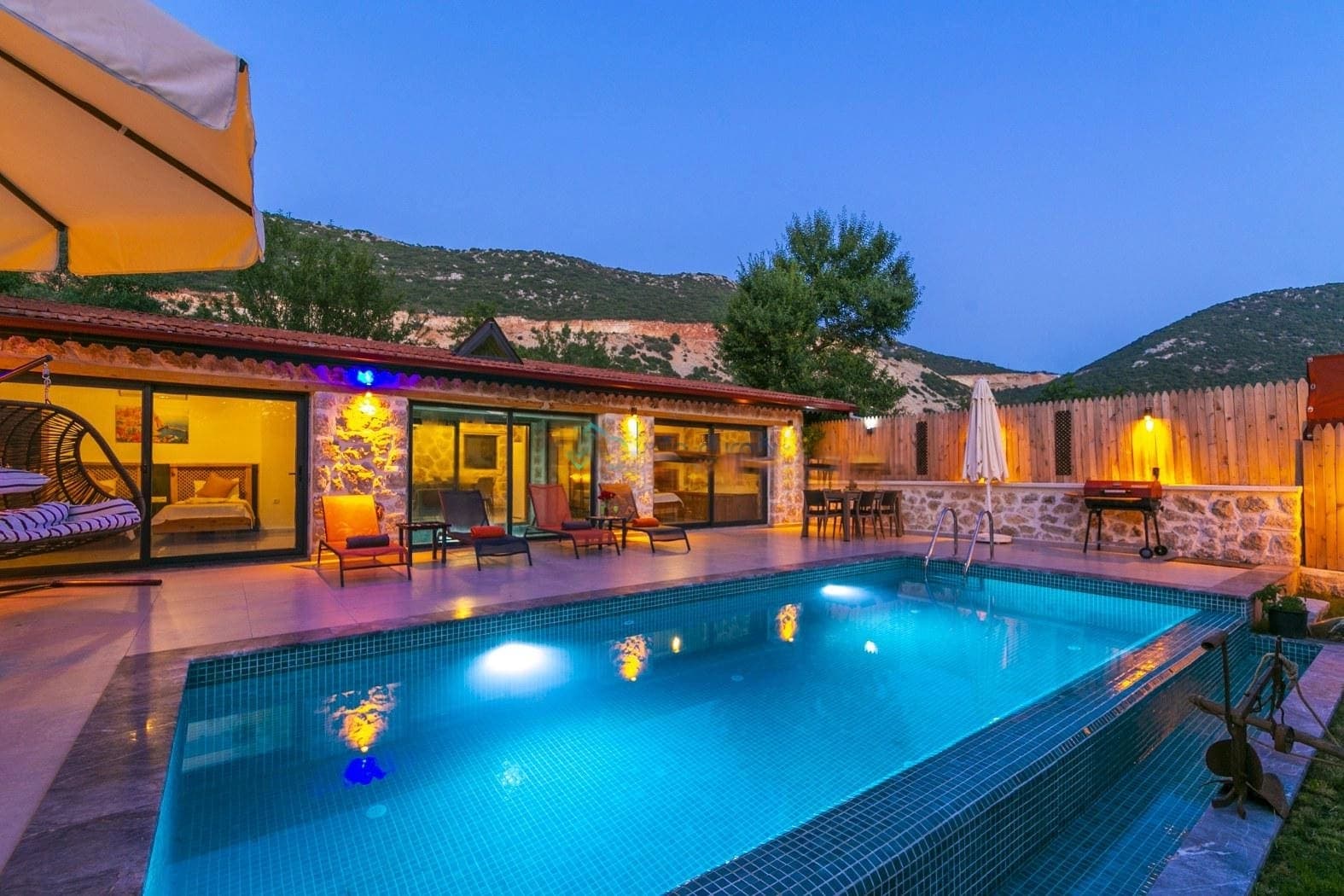 Akşam aydınlatmalı taş villa ve havuzbaşı oturma alanı.