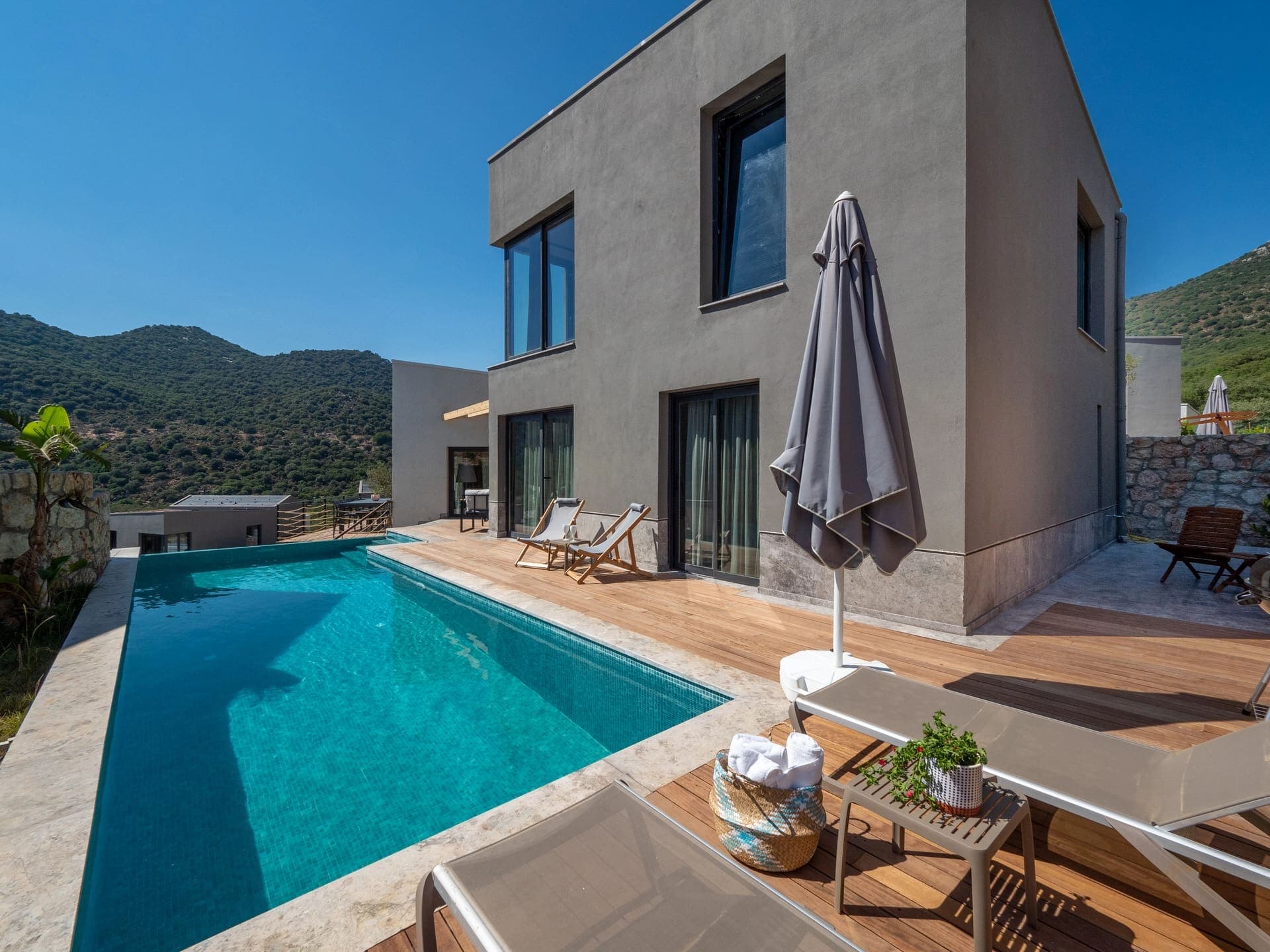 Ahşap teraslı, şezlonglu havuz başı ve gri dış cepheli villa.